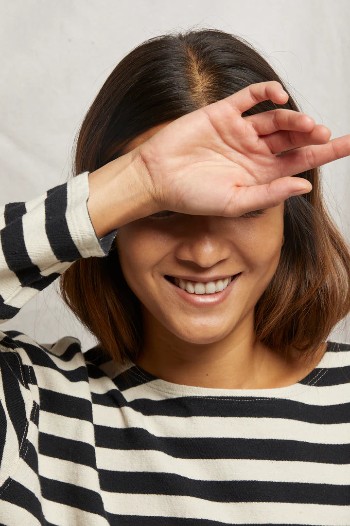 A person wearing the Perfectwhitetee Taylor Long Sleeve Tee, featuring black and white stripes made from recycled cotton, smiles while raising an arm to partially cover their face. Their walnut-shaded, shoulder-length brown hair contrasts with a plain background.