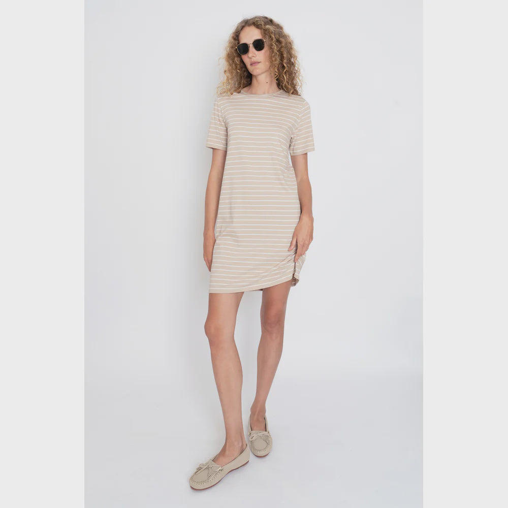 A person wearing Kule's The Tee Dress Sand/White Pinstripe stands against a plain white background. With curly hair and beige moccasins, they exude effortless style.