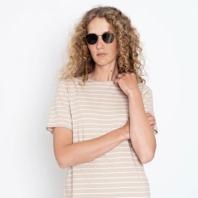 A curly-haired person in sunglasses and Kule's The Tee Dress Sand/White Pinstripe stands confidently against a plain white background. Made from 100% cotton, the dress complements their relaxed pose, with one arm crossed and the other hand on their shoulder.