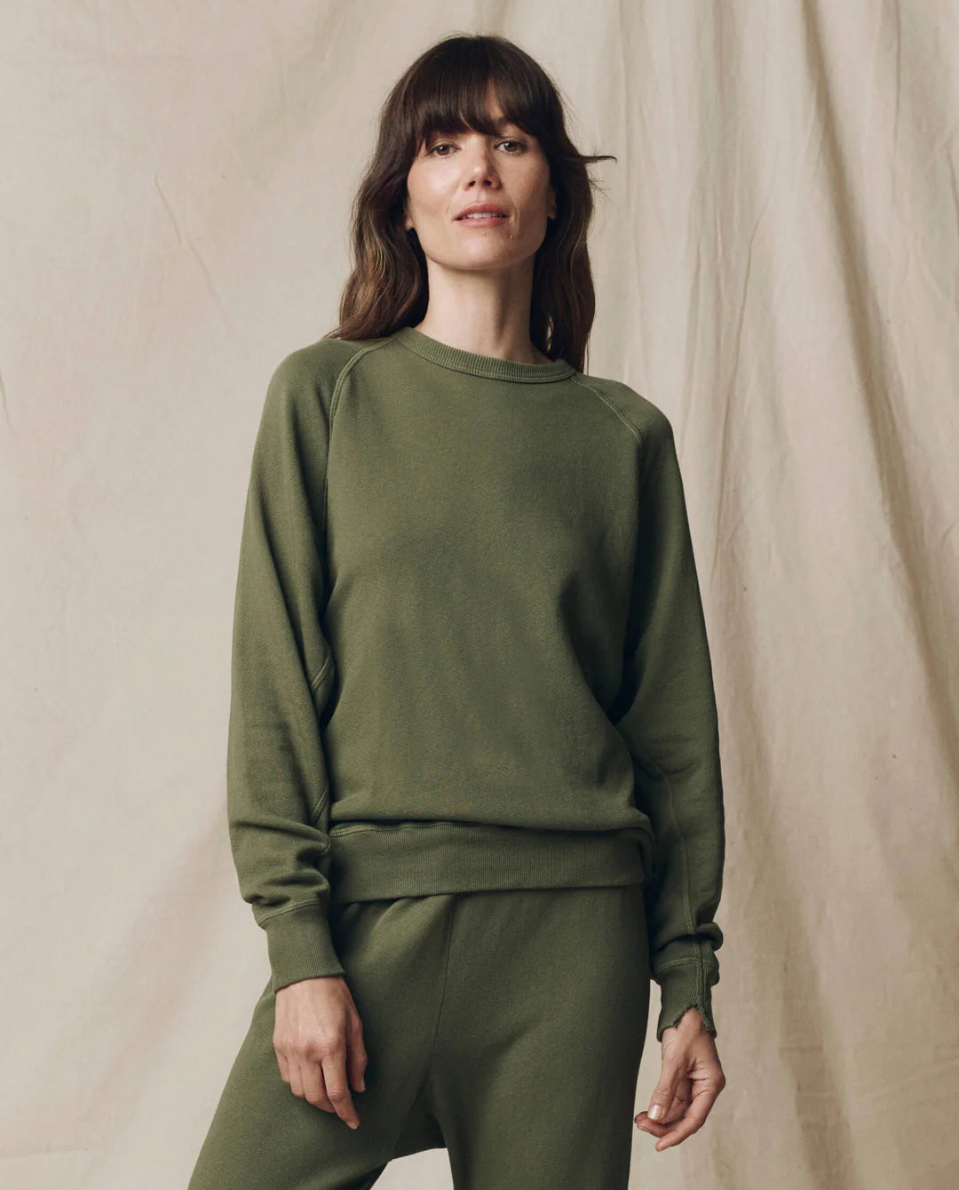 A woman with brown hair, wearing The College Sweatshirt S25 by The Great Inc. and matching pants in olive green, stands smiling gently at the camera before a beige fabric backdrop.