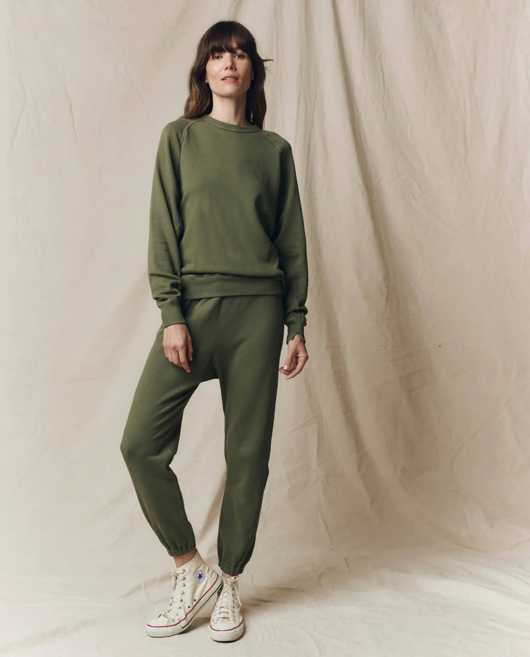 A woman with long brown hair and bangs stands relaxed before a beige fabric backdrop, wearing The Great Inc.'s olive green Stadium Sweatpant and matching sweatshirt, paired with unique, colorful high-top sneakers.