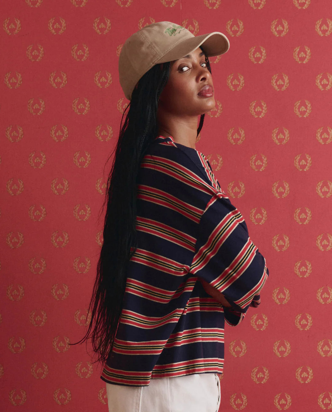 A woman with long black hair, wearing a beige cap and The Field Tee by The Great Inc., featuring navy blue, red, and white stripes, stands before a red patterned backdrop with her arms folded and head turned to the side.