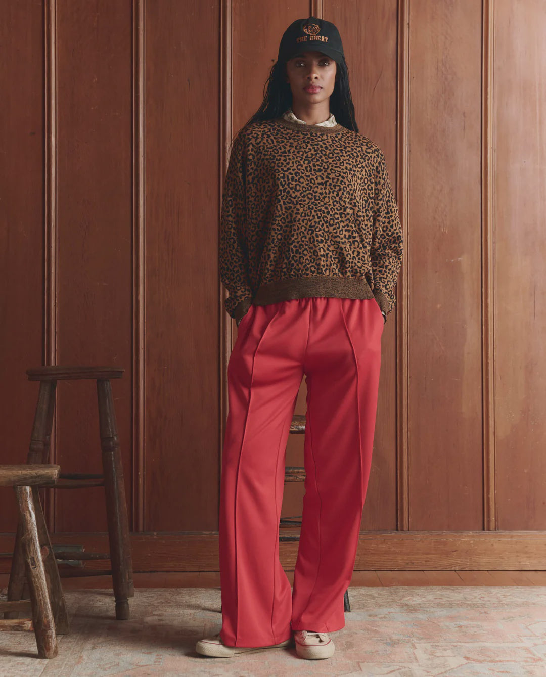 Indoors against wood paneling, a person wears The Great Inc.'s Wide Leg Track Pant in red with an elastic waistband, paired with a black cap, leopard print sweater, and beige shoes. Two wooden stools rest on the rustic wood floor nearby.