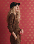 A person with long, curly blonde hair wears The Great Inc.'s The Shrunken Vneck Cardigan in leopard print and a black cap, standing before a red background with gold motifs, one hand in their pocket and a relaxed expression.