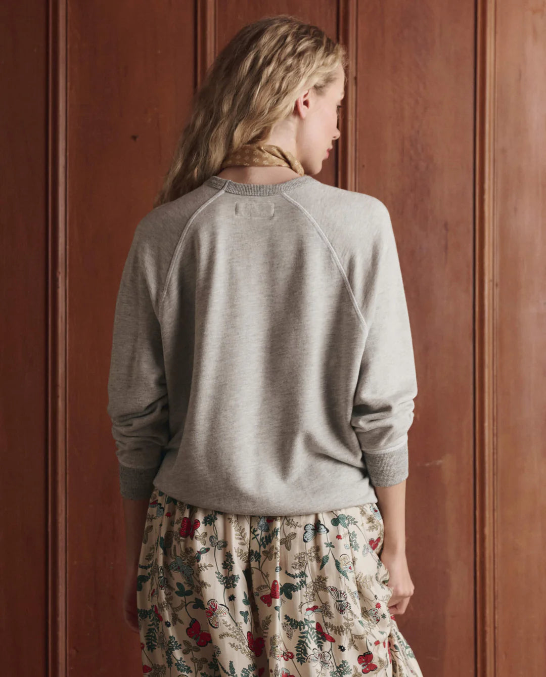 A person with long, wavy blonde hair faces a wooden wall, wearing The College Sweatshirt 2026 by The Great Inc. and a cream floral skirt with colorful botanical patterns—showcasing effortless handcrafted LA style.