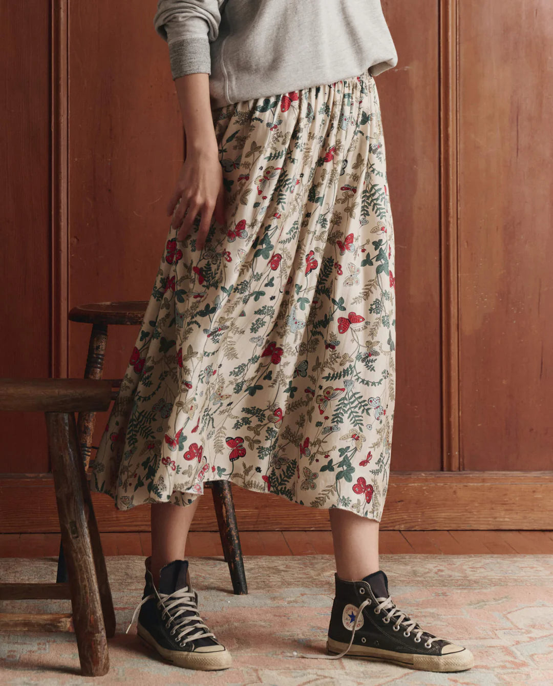 A person wearing The Canary Skirt by The Great Inc.—a cream pull-on skirt with red and green patterns—plus a gray sweatshirt and black high-top sneakers stands indoors beside a wooden stool and wall.