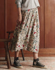 A person wearing The Canary Skirt by The Great Inc.—a cream pull-on skirt with red and green patterns—plus a gray sweatshirt and black high-top sneakers stands indoors beside a wooden stool and wall.