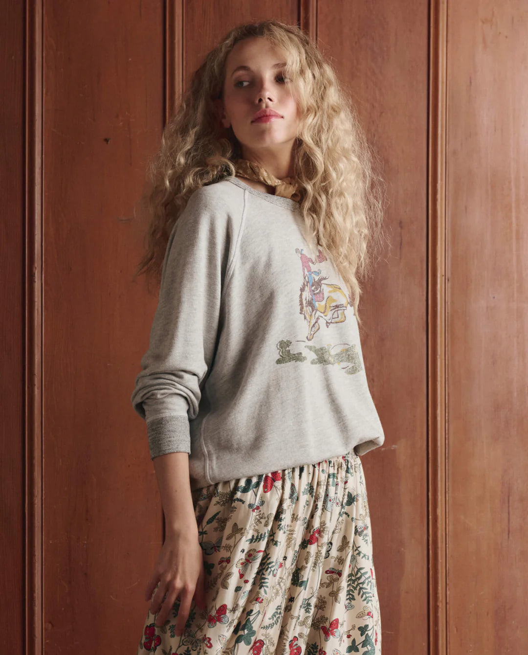 A woman with long, curly blonde hair wears The College Sweatshirt 2026 by The Great Inc. over a floral skirt, standing before a wooden paneled wall and glancing slightly over her shoulder.