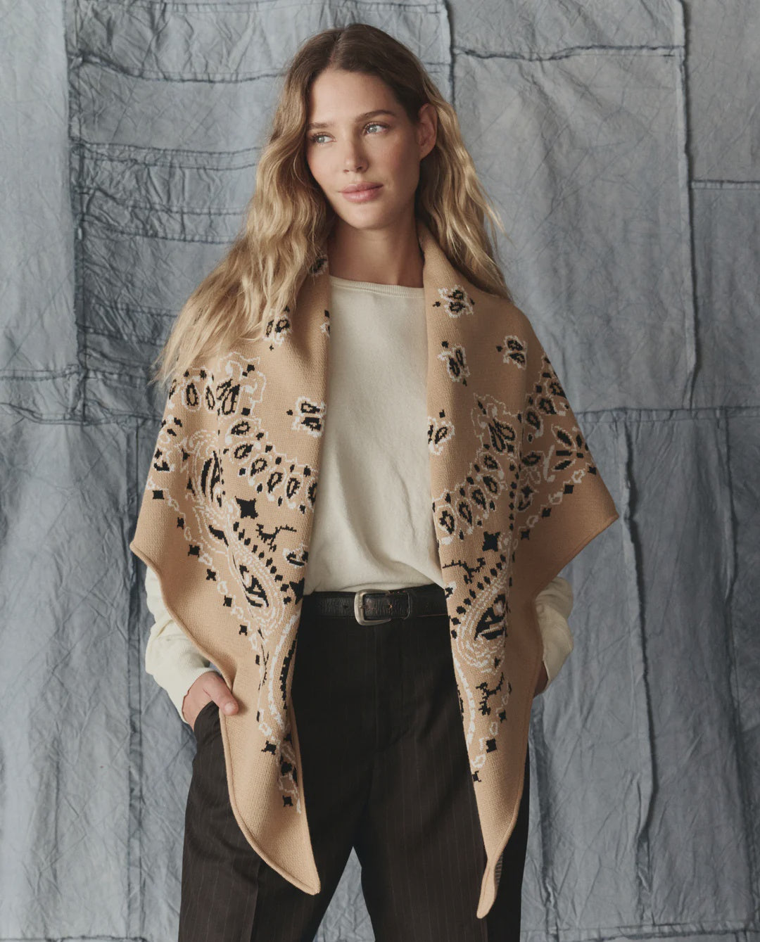 A woman with long wavy hair stands against a textured blue-grey background, wearing a cream shirt, dark trousers, and The Great Inc.'s The Big Bandana Scarf styled as an oversized wrap draped over her shoulders.