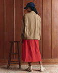 A person stands indoors facing away, wearing The Folsom Jacket by The Great Inc. over a red midi skirt, beige shoes, and a navy cap. A wooden stool is beside them, set against wood-paneled walls for a nostalgic atmosphere.
