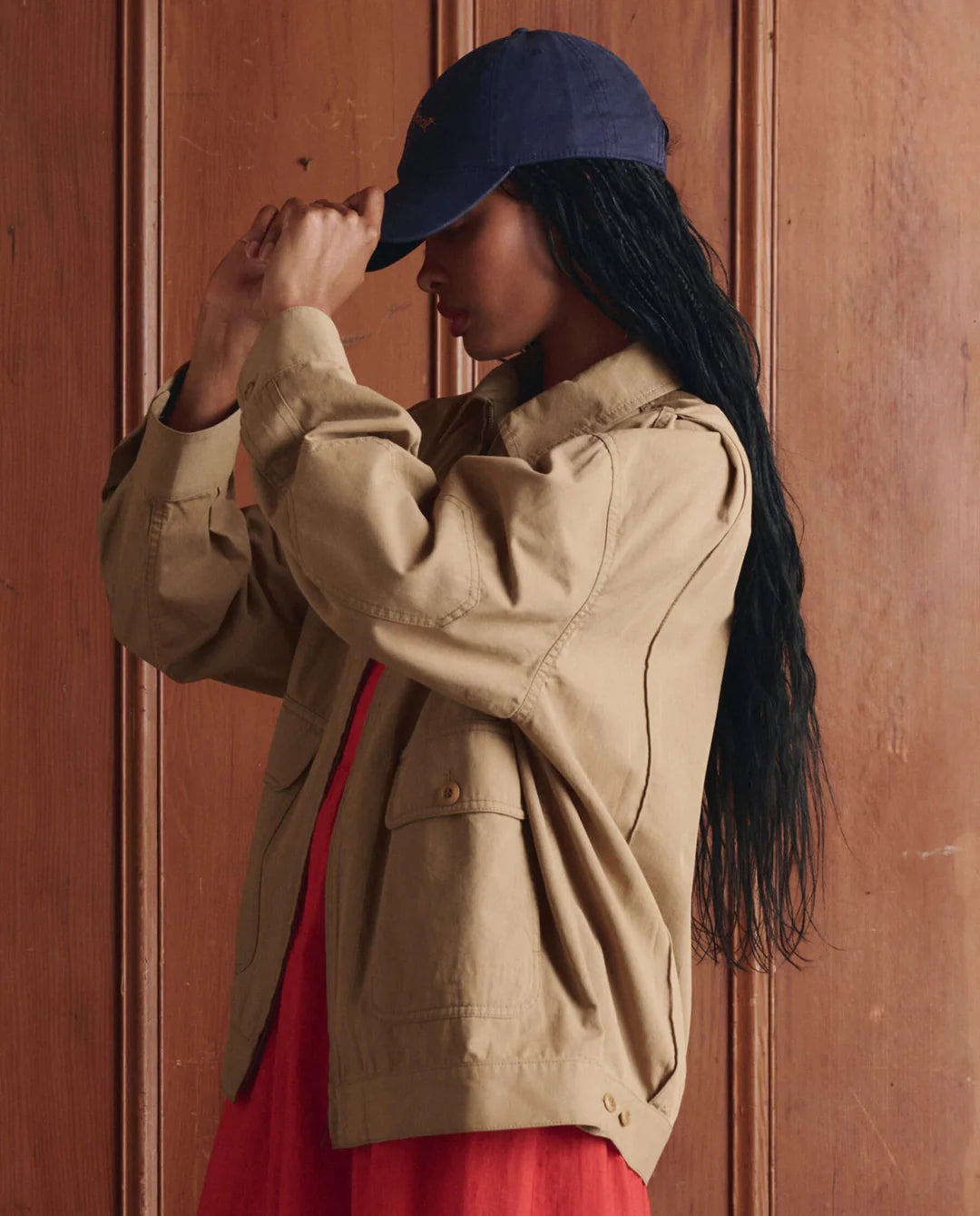 A person with long dark hair wears The Folsom Jacket by The Great Inc. in tan cotton twill, paired with a red-orange dress and navy cap, standing sideways against a wooden wall and adjusting the cap for a 1940s workwear vibe.