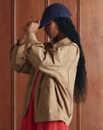 A person with long dark hair wears The Folsom Jacket by The Great Inc. in tan cotton twill, paired with a red-orange dress and navy cap, standing sideways against a wooden wall and adjusting the cap for a 1940s workwear vibe.