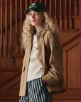 A person with long curly blonde hair wears a green cotton twill baseball cap, graphic t-shirt, blue striped pants, and The Folsom Jacket by The Great Inc., while standing in front of wooden stairs.