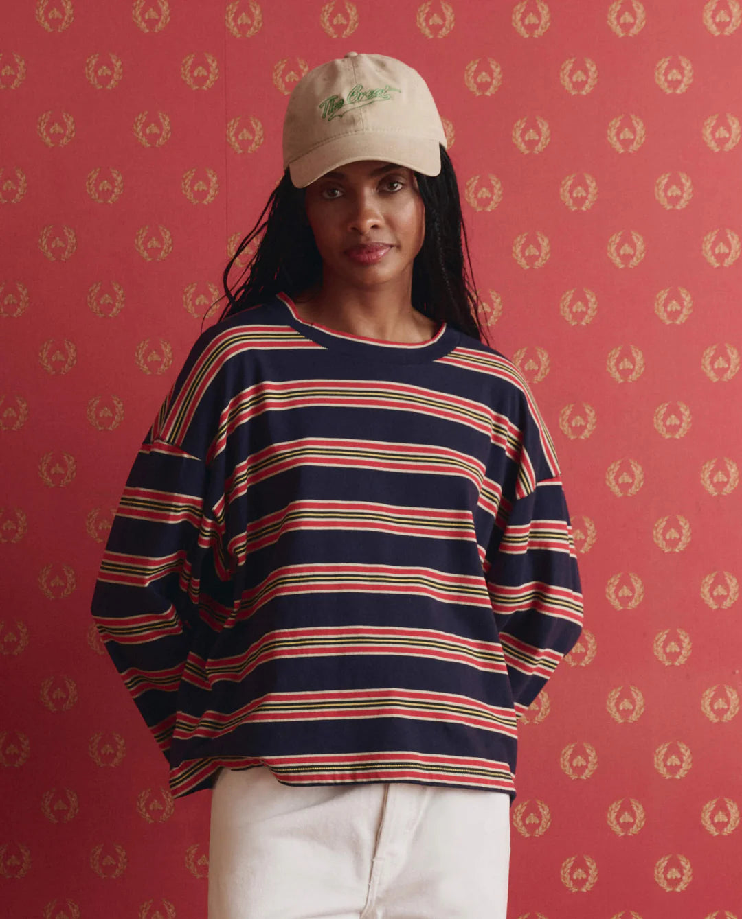 A person wearing a beige cap, The Great Inc. The Field Tee in navy blue stripes, and white pants stands against a red patterned background, looking at the camera.