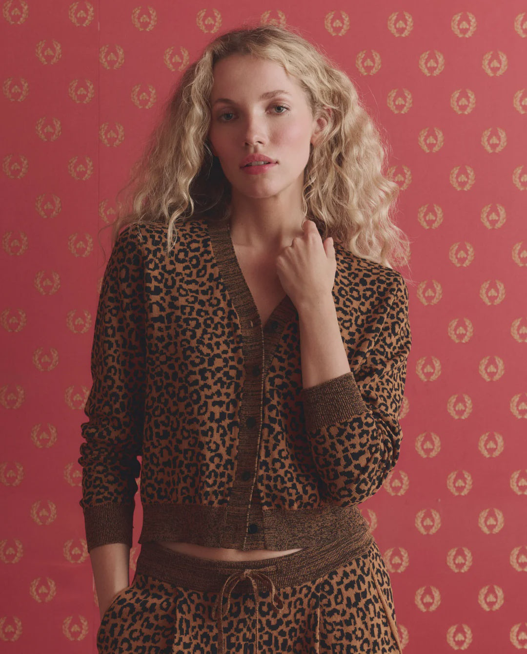 A woman with long curly blonde hair wears The Great Inc.'s The Shrunken Vneck Cardigan and matching pants, standing against a red patterned background with gold motifs. She faces forward, one hand touching her collar.