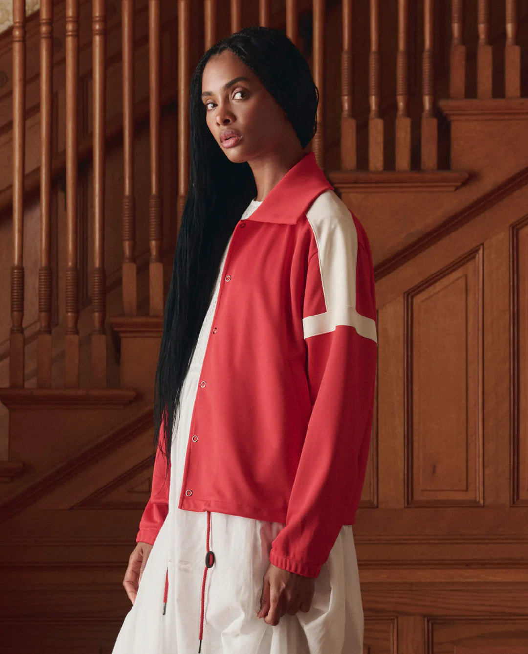 A woman with long black hair wears The Trainer Jacket by The Great Inc., featuring vintage styling and white stripes, over a white dress, standing indoors before wooden stairs and paneling, gazing slightly to the side.