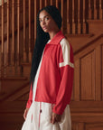 A woman with long black hair wears The Trainer Jacket by The Great Inc., featuring vintage styling and white stripes, over a white dress, standing indoors before wooden stairs and paneling, gazing slightly to the side.