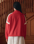 A person with long dark hair stands facing away, wearing The Trainer Jacket by The Great Inc.—a vintage sport jacket with white stripes—over a white dress, in front of wooden stairs and paneling.
