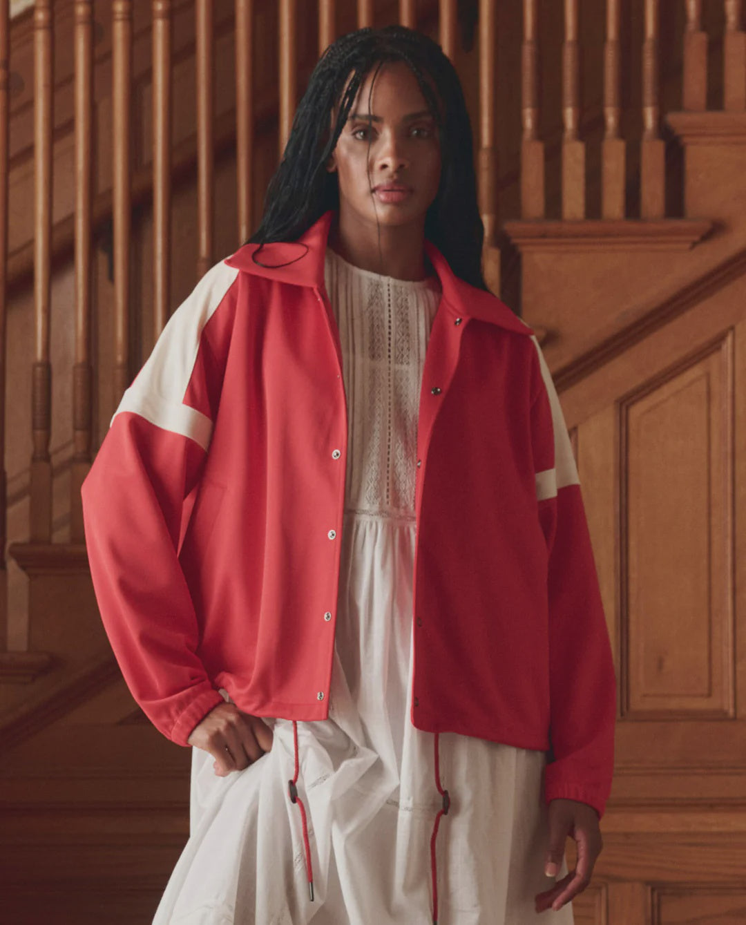A person with long dark hair stands indoors by a wooden staircase, wearing The Trainer Jacket by The Great Inc. with white stripes over a white dress.