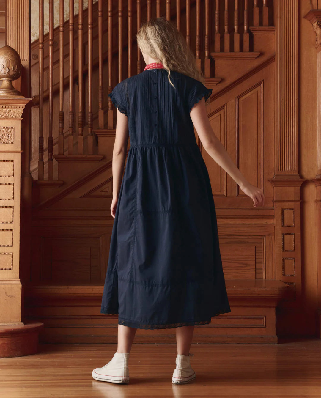 A person with long blonde hair, wearing The Great Inc. Sonnett Dress in navy blue cotton slub and white sneakers, stands with their back to the camera in front of a wooden staircase.
