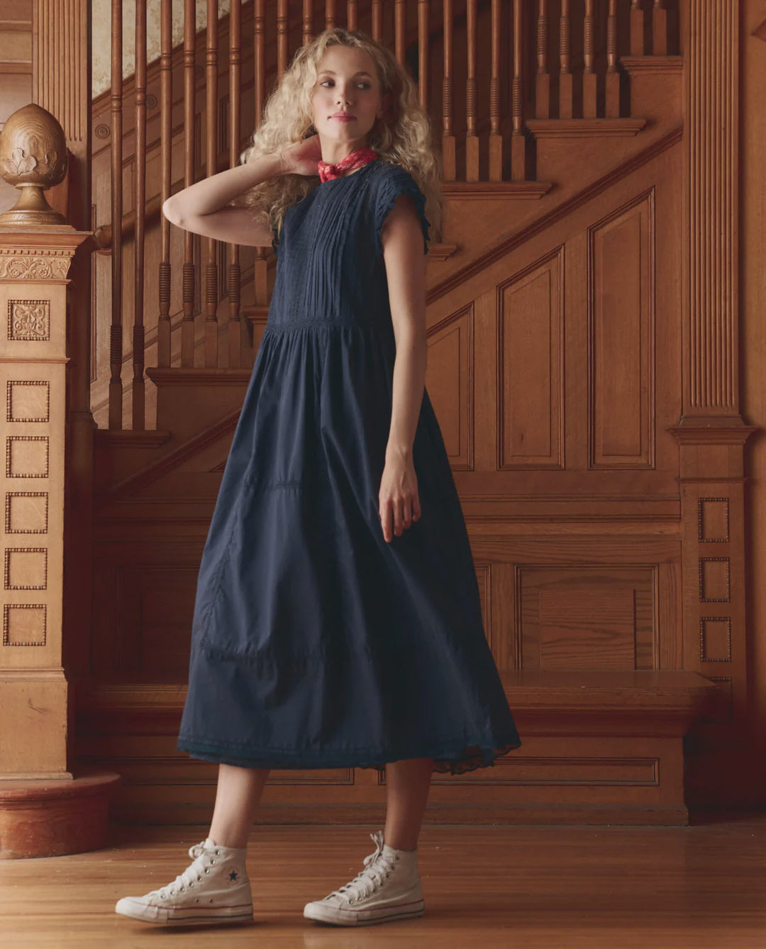 A woman with wavy blonde hair, wearing The Great Inc. Sonnett Dress in navy blue lace, white high-top sneakers, and a red scarf stands before an ornately carved wooden staircase.