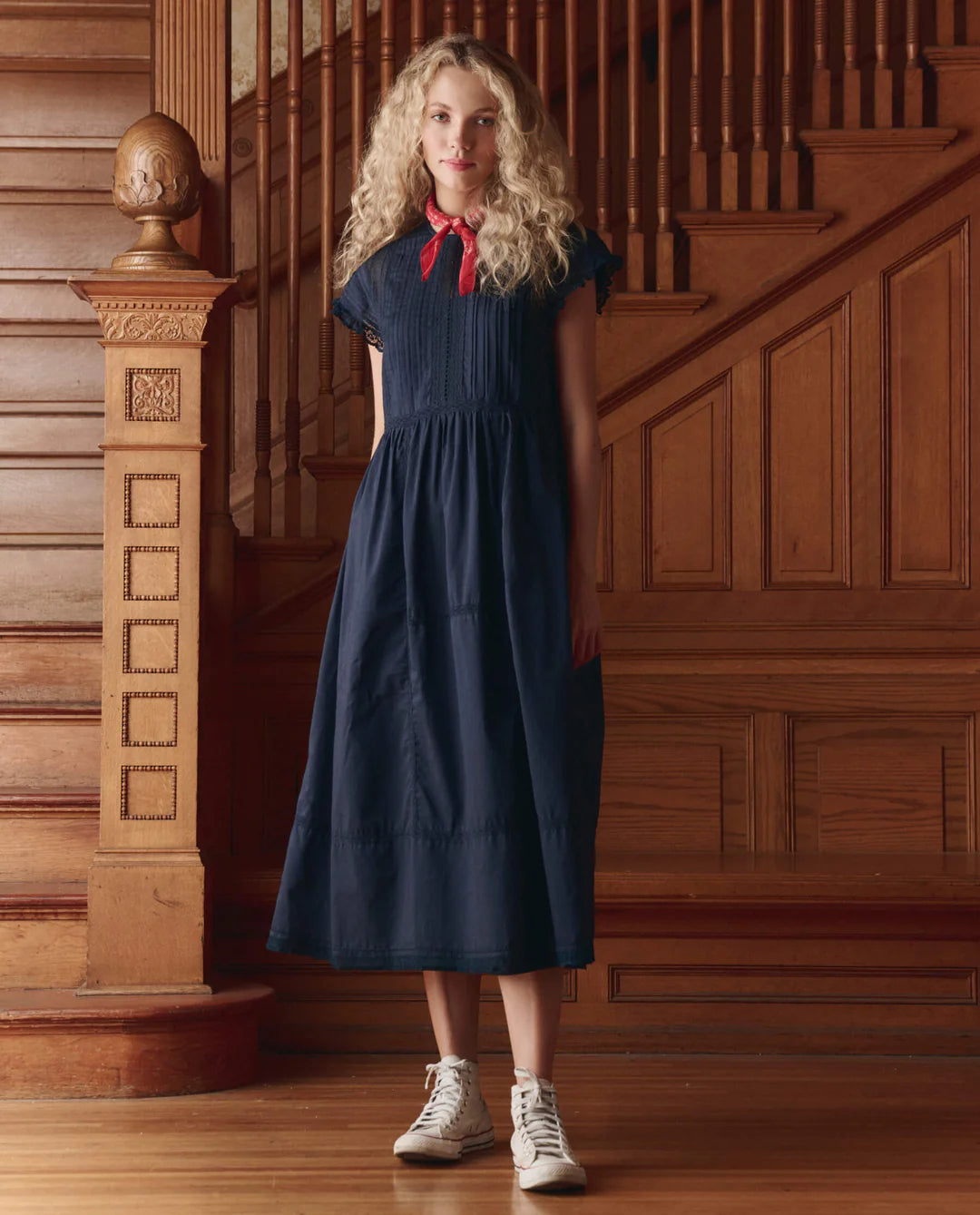 A woman with curly blonde hair, wearing The Great Inc. Sonnett Dress in navy blue cotton slub, a red neck scarf, and white sneakers, stands before a wooden staircase and paneled wall.
