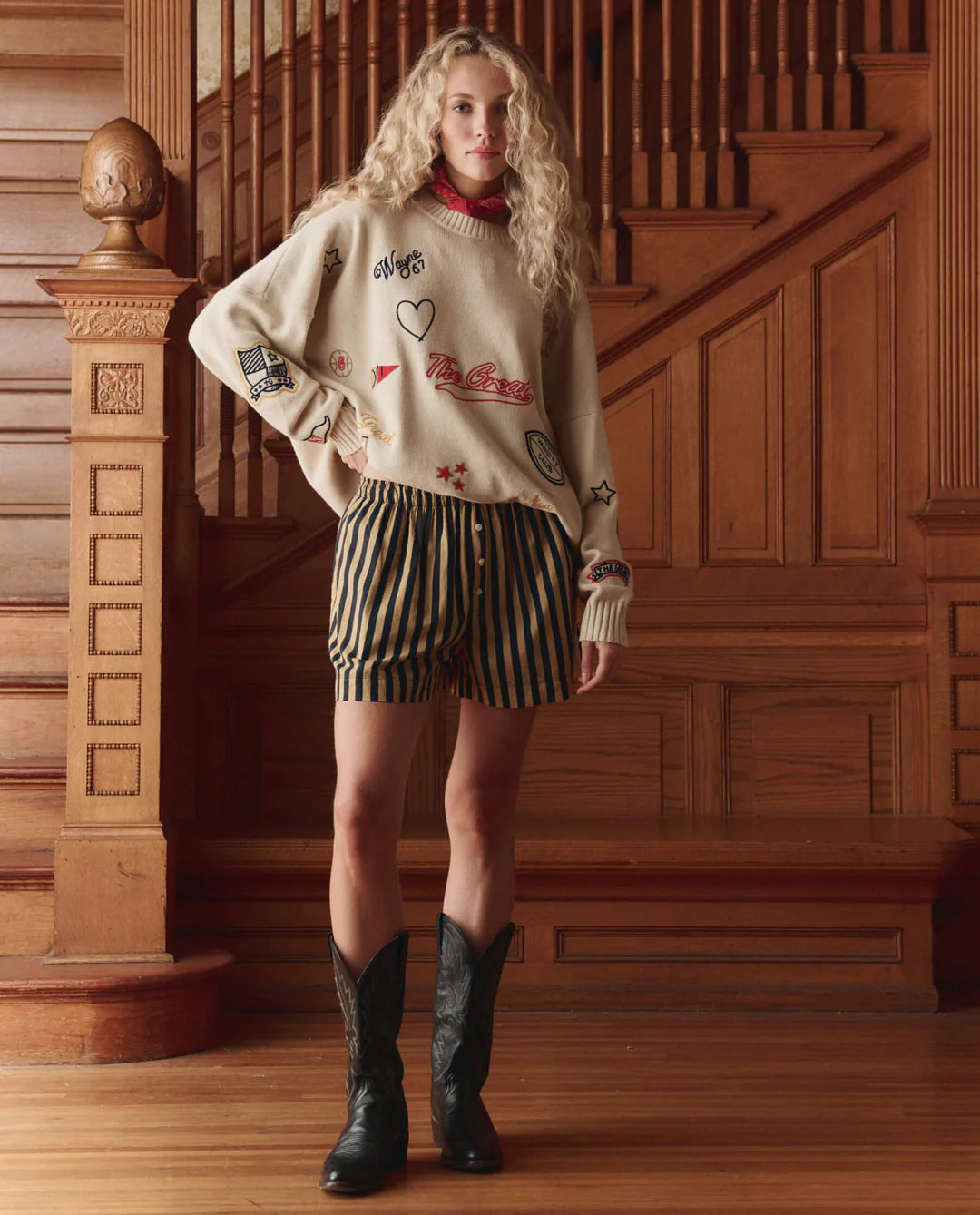A person with long curly blonde hair stands by a wooden staircase indoors, wearing The Embroidered Oversize Crew by The Great Inc., striped shorts, and black cowboy boots.