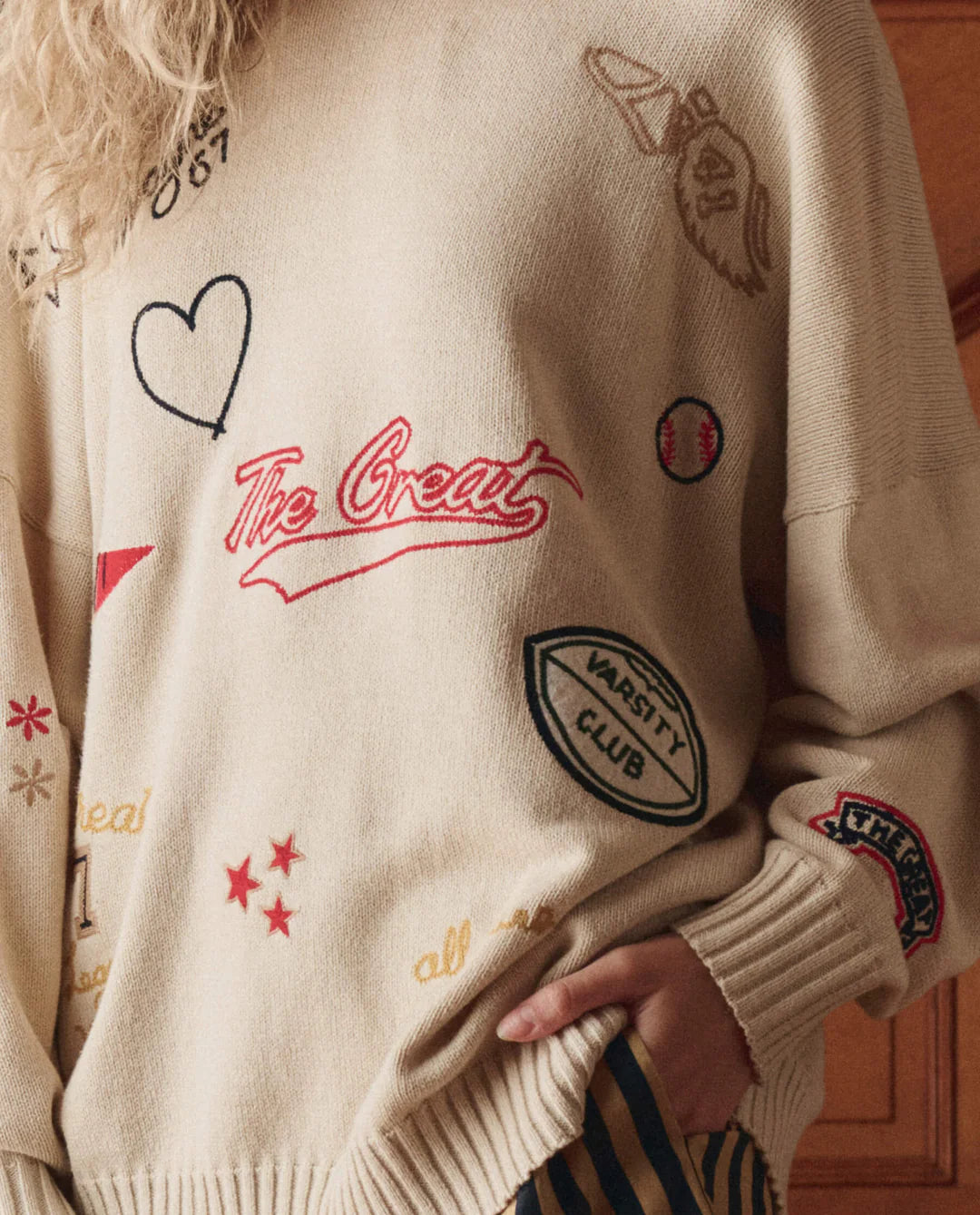 A person wears The Great Inc.'s Embroidered Oversize Crew, a beige cotton-blend sweater styled like a vintage varsity jacket with heart, star, "The Great," and football patches. Their blond hair is partly visible as they rest one hand in their pocket.