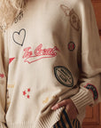 A person wears The Great Inc.'s Embroidered Oversize Crew, a beige cotton-blend sweater styled like a vintage varsity jacket with heart, star, "The Great," and football patches. Their blond hair is partly visible as they rest one hand in their pocket.