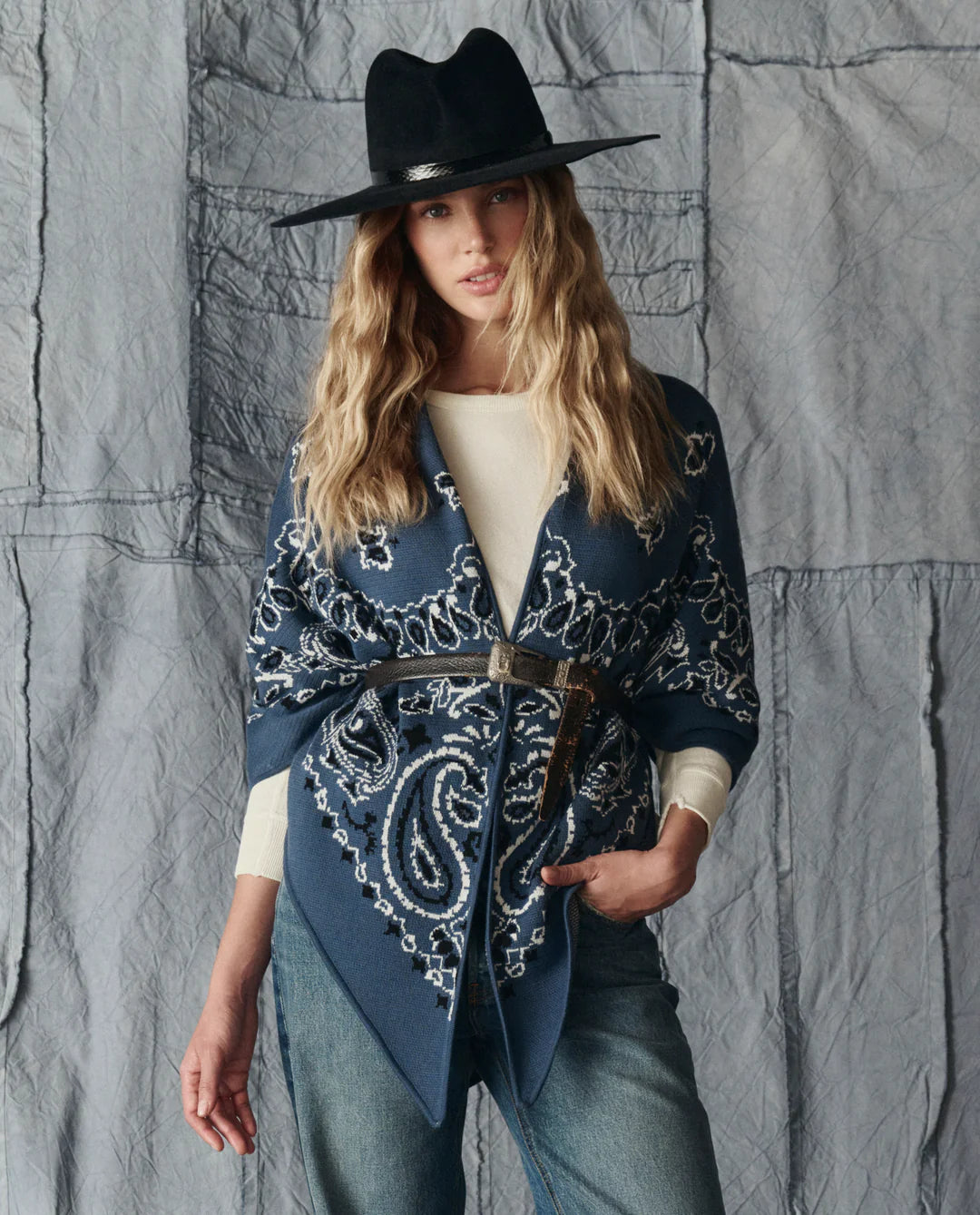 A woman stands before a textured gray backdrop, wearing The Great Inc.'s The Big Bandana Scarf belted at the waist over a cream long-sleeve top, blue jeans, and a wide-brimmed black hat.