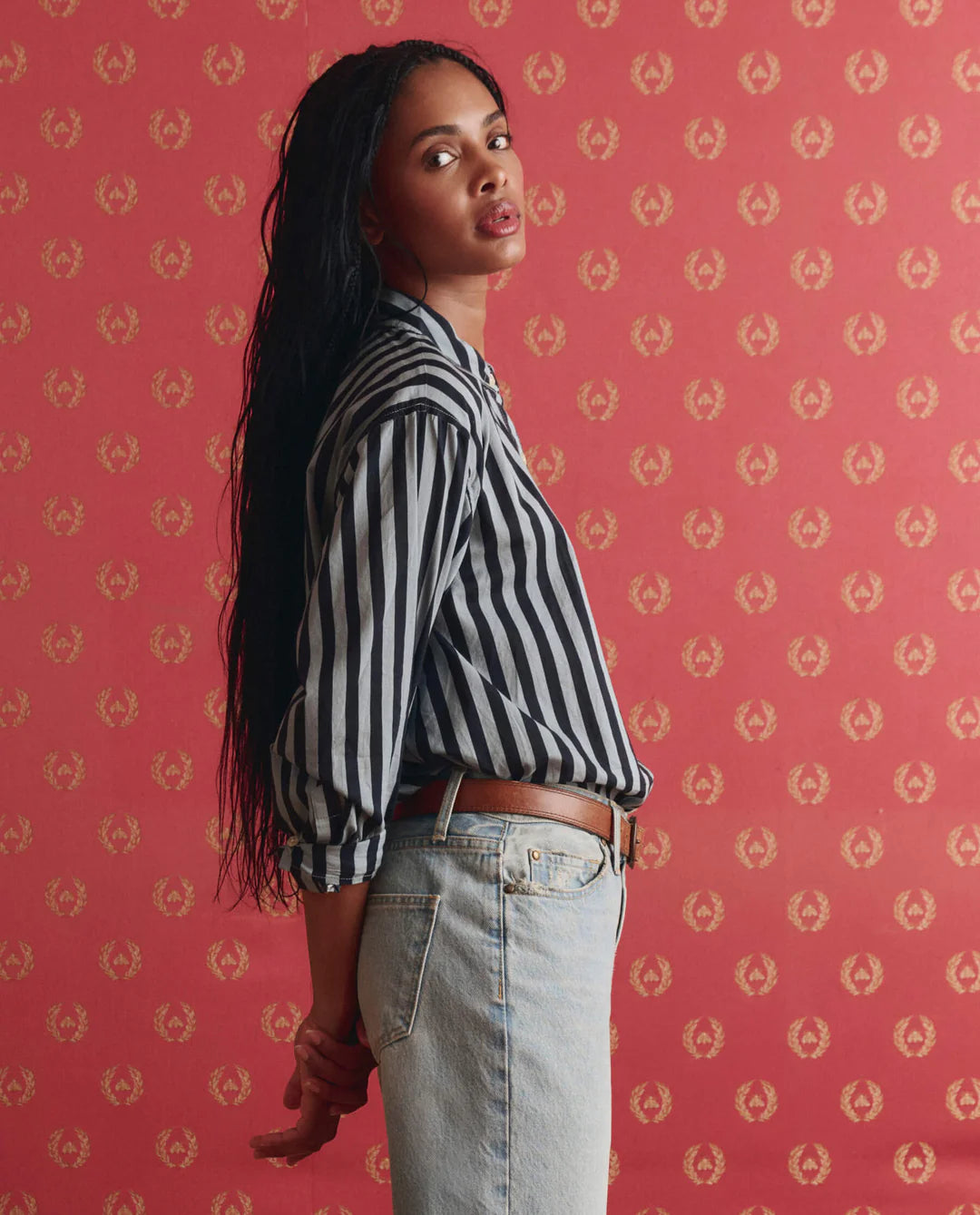 A woman with long dark hair stands sideways against red patterned wallpaper, wearing The Daily Button Up by The Great Inc.—an oversized black-and-white striped shirt—and light blue jeans, gazing at the camera with a relaxed expression.