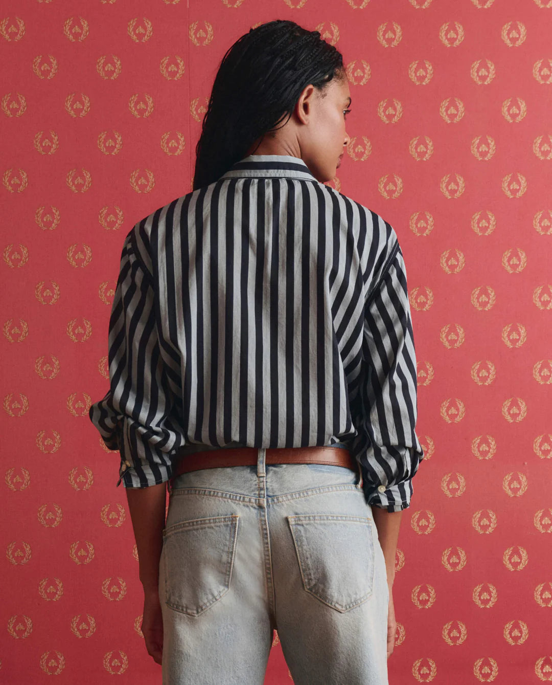 A person with long dark hair, seen from behind, wears The Daily Button Up by The Great Inc., featuring black-and-white stripes, paired with light blue jeans, standing before red wallpaper with a gold pattern.