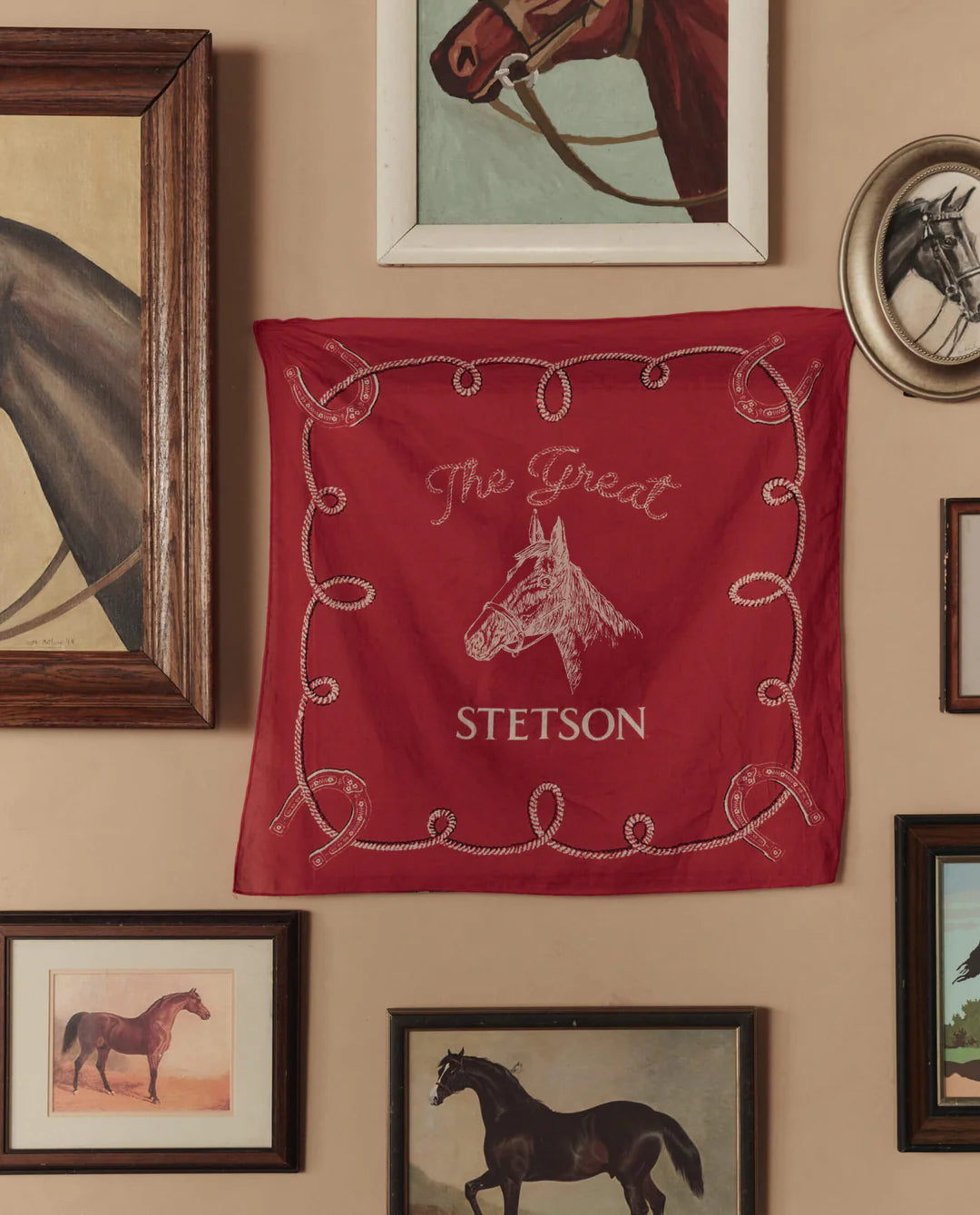 The Stetson Western Bandana by The Great Inc., featuring "The Great Stetson" and a horse head design, is displayed on a beige wall among framed horse paintings, adding vintage charm to any handkerchief-loving space.