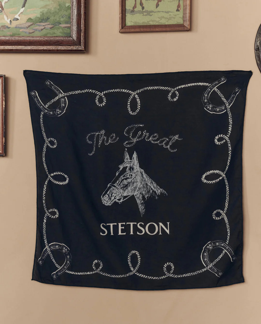 The Stetson Western Bandana by The Great Inc., featuring a black design with a white horse head, "The Great STETSON" text, and a rope with horseshoes border, hangs on the wall beneath framed paintings.