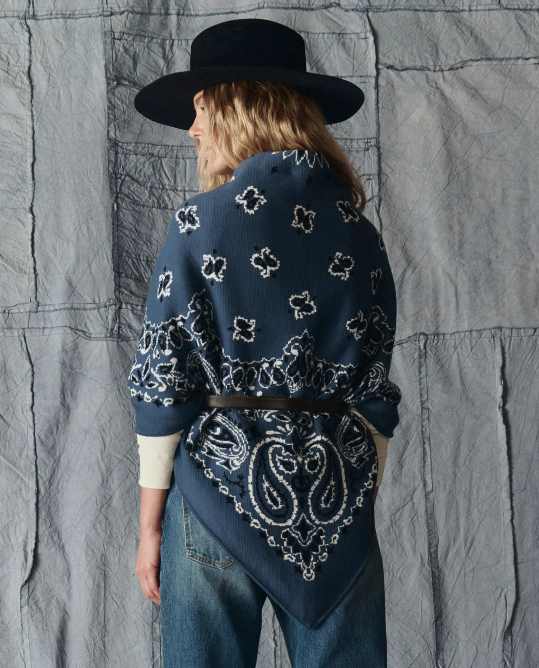 A person stands with their back to the camera, wearing a wide-brimmed black hat, cream shirt, blue jeans, and The Big Bandana Scarf by The Great Inc., belted at the waist, against a gray fabric backdrop.