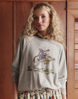 A woman with long, curly blonde hair wears The College Sweatshirt 2026 by The Great Inc.—an oversized vintage washed terry style with a cowboy horse graphic—paired with a floral skirt, standing indoors against wooden paneling.