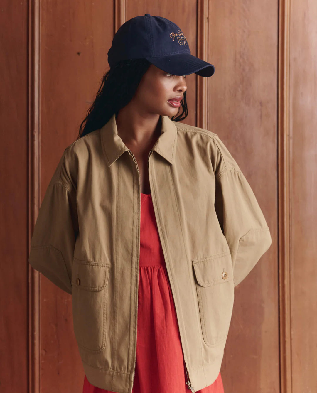 A woman stands before a wooden paneled wall, hands behind her back, wearing The Great Inc.'s The Folsom Jacket over a red dress and a navy blue baseball cap, looking to the side.