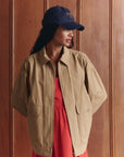 A woman stands before a wooden paneled wall, hands behind her back, wearing The Great Inc.'s The Folsom Jacket over a red dress and a navy blue baseball cap, looking to the side.