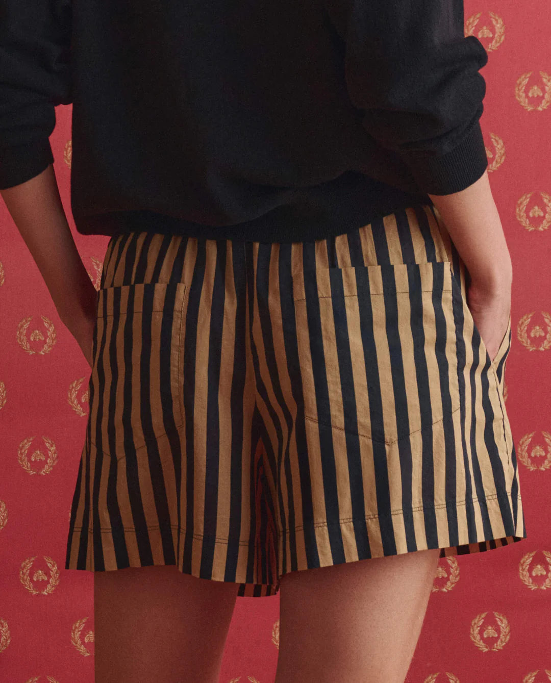 A person models The Great Inc.'s The Boxer Short in navy and tan vertical stripes with a black top, standing against a red backdrop adorned with small gold laurel wreaths.