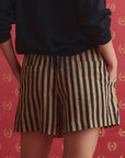 A person models The Great Inc.'s The Boxer Short in navy and tan vertical stripes with a black top, standing against a red backdrop adorned with small gold laurel wreaths.