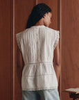 A woman with long dark hair stands facing away, wearing The Sonnet Top by The Great Inc., a Victorian-inspired blouse with lace detailing, pleats, and back buttons. She is in front of a wooden paneled wall with part of an open book visible.