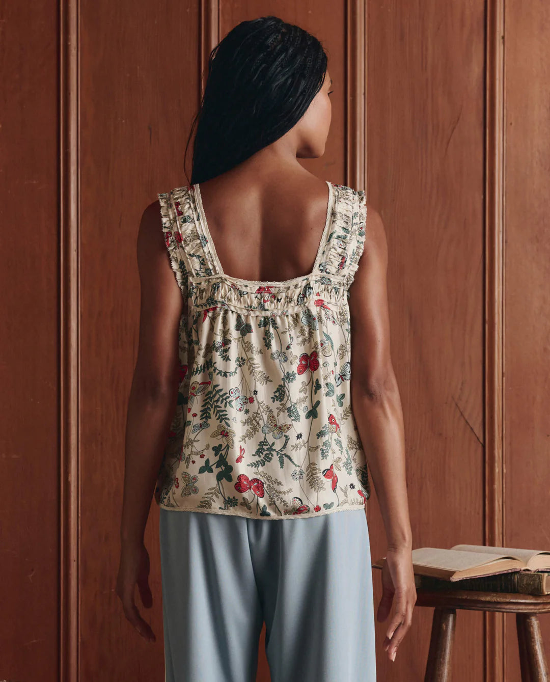 A woman faces a wooden wall, wearing The Great Inc.'s lightweight cotton Victorian Pleat Tank with ruffle details and light blue pants. An open book sits on a small wooden table next to her.