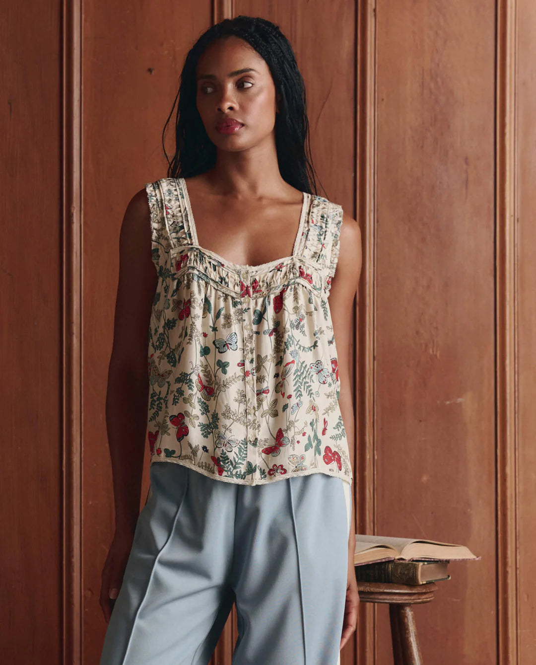 A woman stands indoors by a wooden wall, wearing The Great Inc.'s Victorian Pleat Tank in vintage rose print with ruffled straps and light blue pants. An open book sits on a small stool beside her.