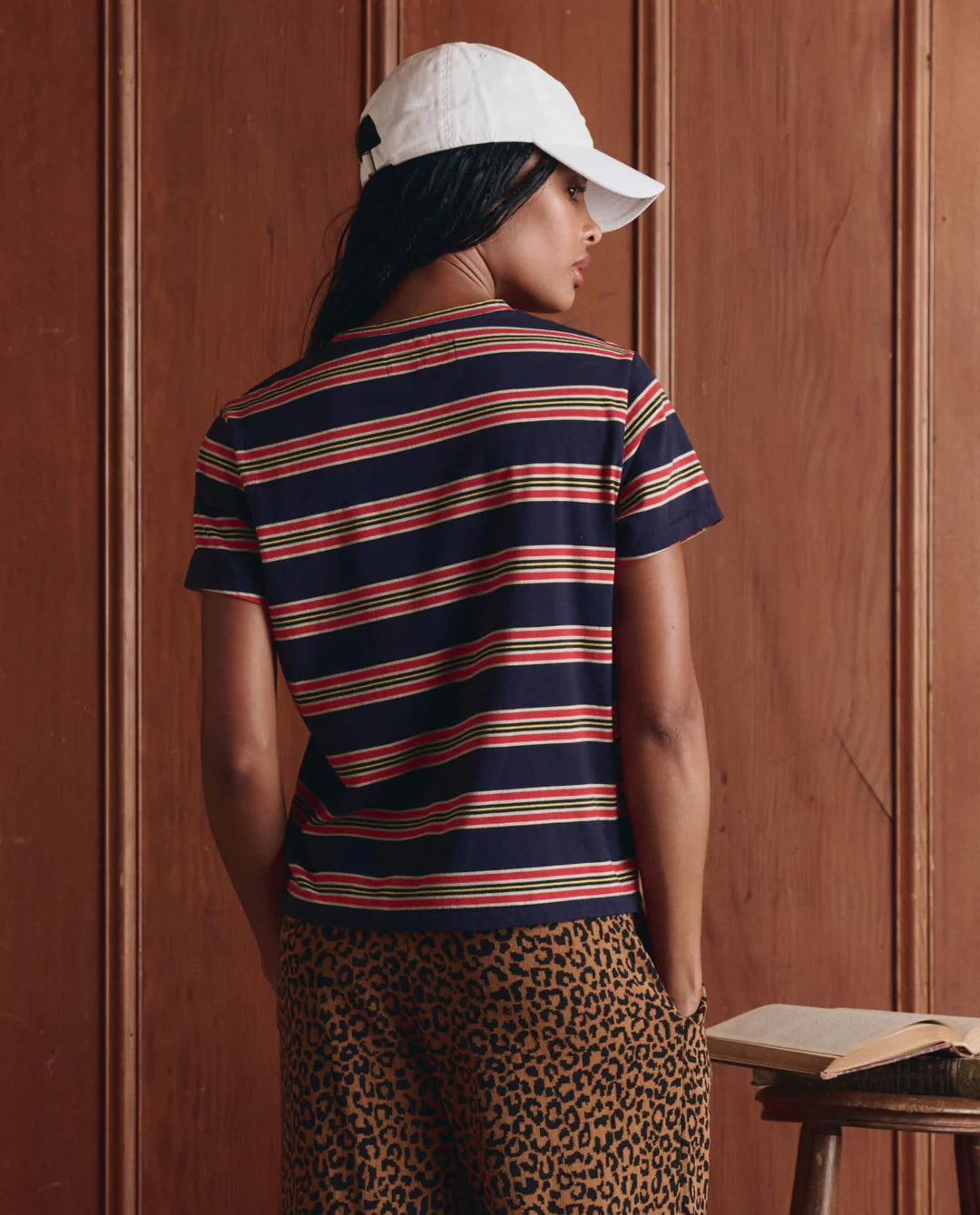 Wearing The Little Tee by The Great Inc. in navy blue stripes, paired with leopard print pants and a white cap, a person stands with their back to the camera before a wooden wall. An open book rests on a table nearby.