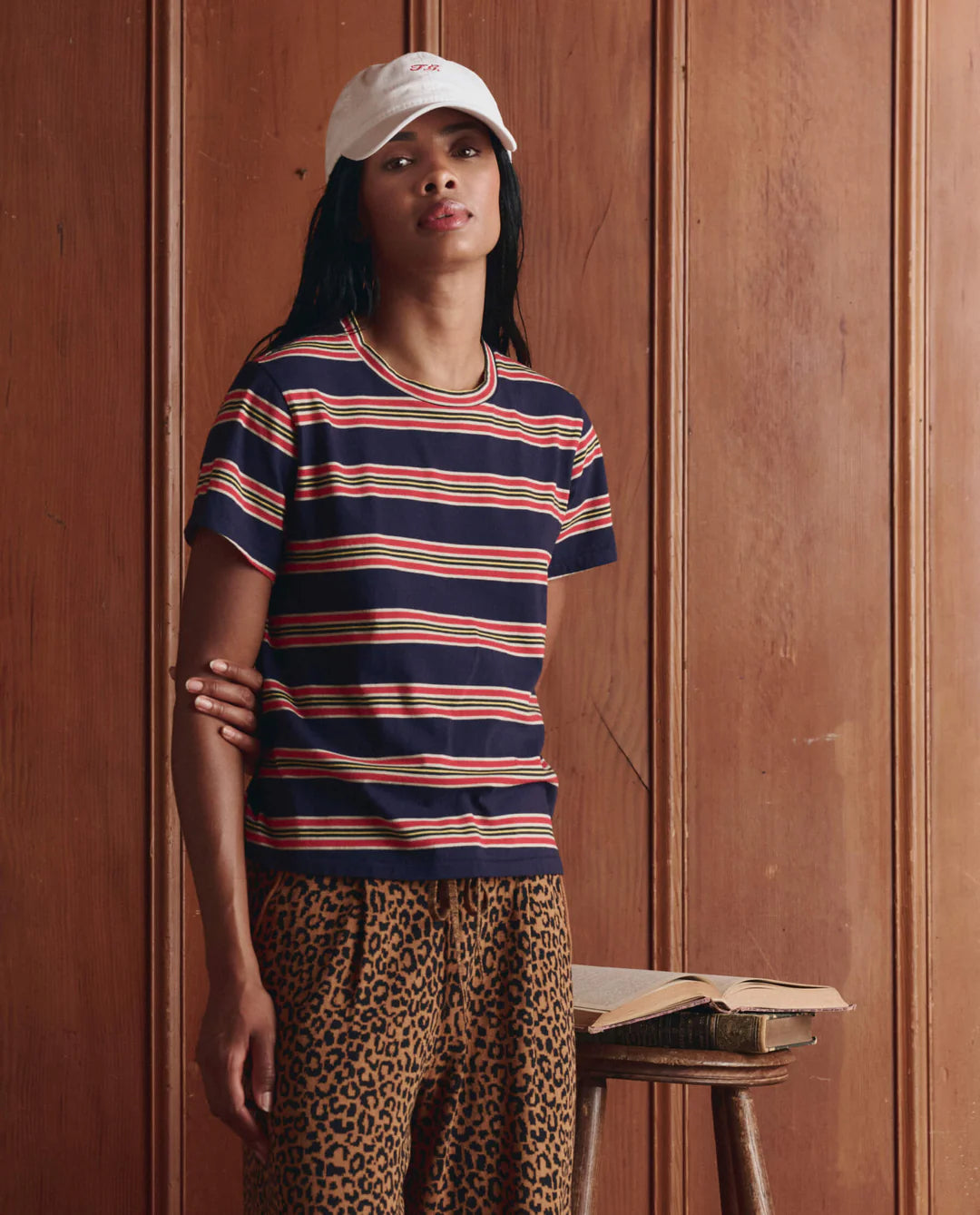 A person in The Little Tee by The Great Inc., a white cap, and leopard print pants stands indoors beside a stool with an open book, in front of wooden paneled walls.