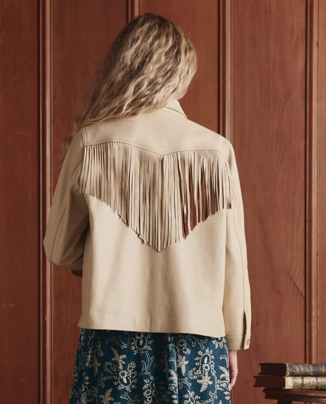 A person with long, wavy blonde hair is seen from behind wearing The Leather Fringe Jacket by The Great Inc., featuring vintage-inspired fringe details across the back and shoulders, in front of wooden paneling and stacked books.