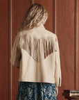 A person with long, wavy blonde hair is seen from behind wearing The Leather Fringe Jacket by The Great Inc., featuring vintage-inspired fringe details across the back and shoulders, in front of wooden paneling and stacked books.