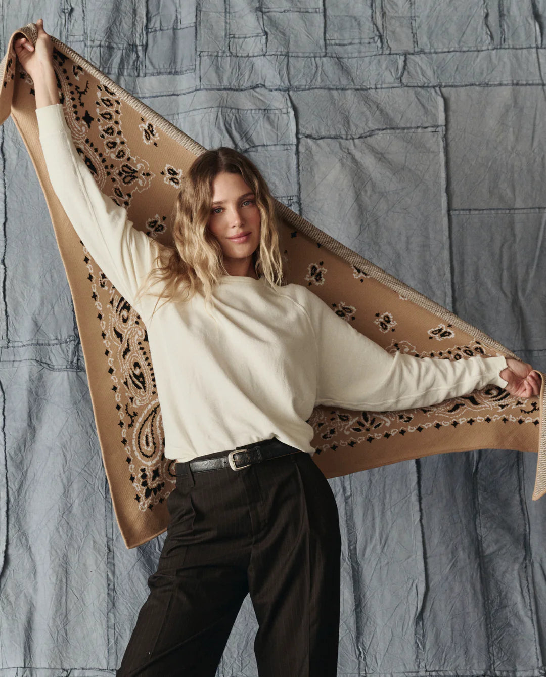 A woman with wavy blonde hair, in a cream sweater and dark pants, displays The Great Inc.'s Big Bandana Scarf—with intricate patterns—against a textured gray backdrop.