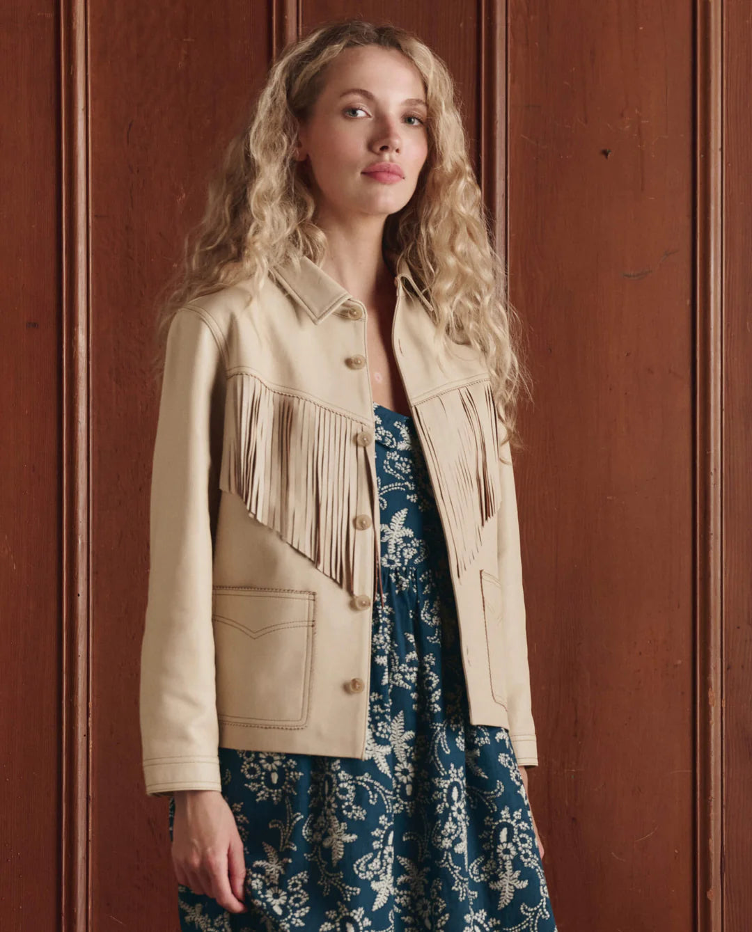 A woman with long curly blonde hair models The Leather Fringe Jacket by The Great Inc. over a blue and white floral dress, standing before a wooden paneled wall.