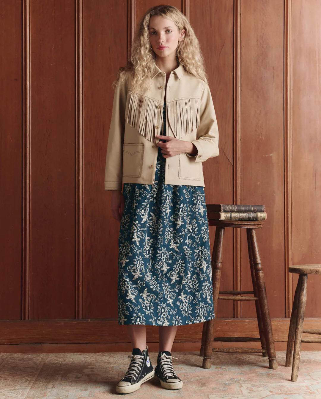 A woman with long, wavy blonde hair stands indoors against wood paneling, wearing The Great Inc. Leather Fringe Jacket over a blue floral midi dress and black high-top sneakers, beside a small stool stacked with books.